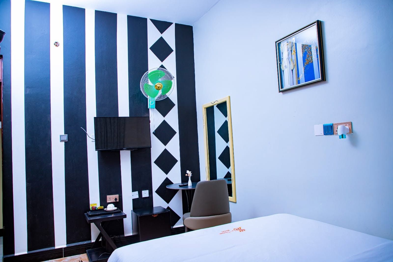 A hotel room with a bed, a desk, a chair, a TV, a fan, and a painting on the wall.