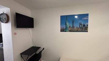 Una habitación con una cama, un escritorio con una silla, un televisor montado en la pared y un cartel con una vista urbana.