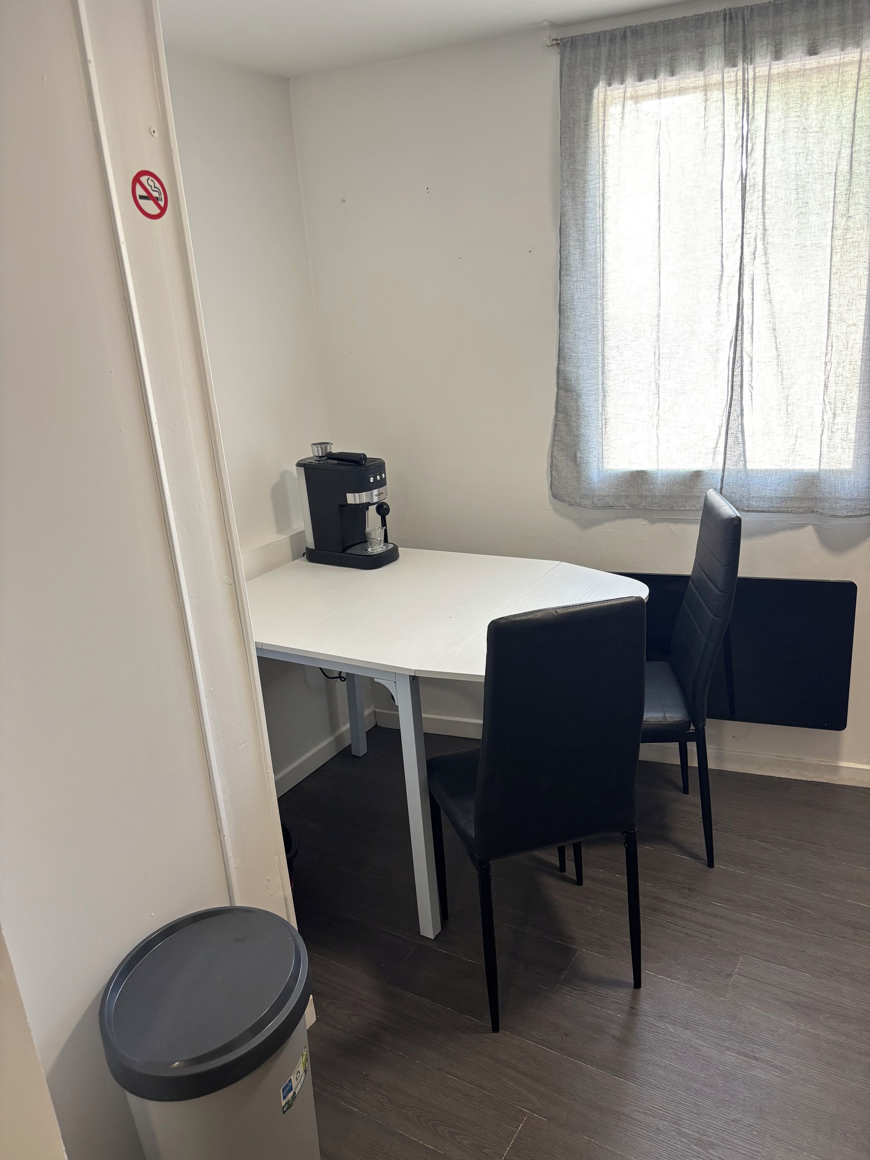 Una stanza con un tavolo bianco, sedie nere, una macchina per caffè, un cestino e una finestra con tenda.