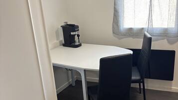 Una stanza con un tavolo bianco, sedie nere, una macchina per caffè, un cestino e una finestra con tenda.