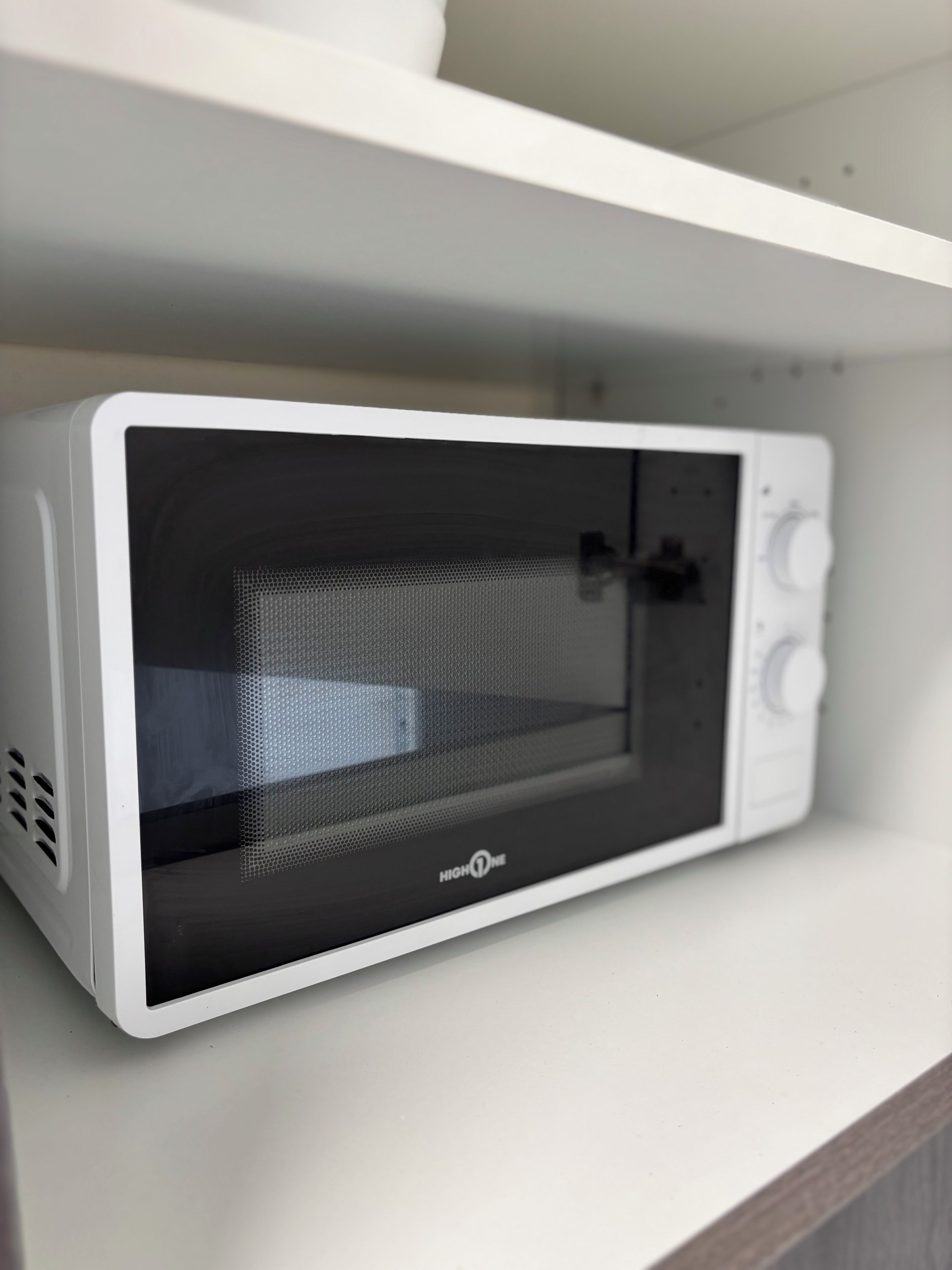 A white microwave oven with a mesh door and control knobs on the right side, placed on a white shelf.