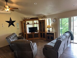 A living room with leather sofas, a television, wooden cabinets, and a ceiling fan. - Charming 3-Bdrm home overlooking the Majestic Coos River with AC. (Coos Bay)