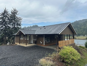 A two-story house with a covered porch, a gravel driveway, and a view of a lake. - Charming 3-Bdrm home overlooking the Majestic Coos River with AC. (Coos Bay)