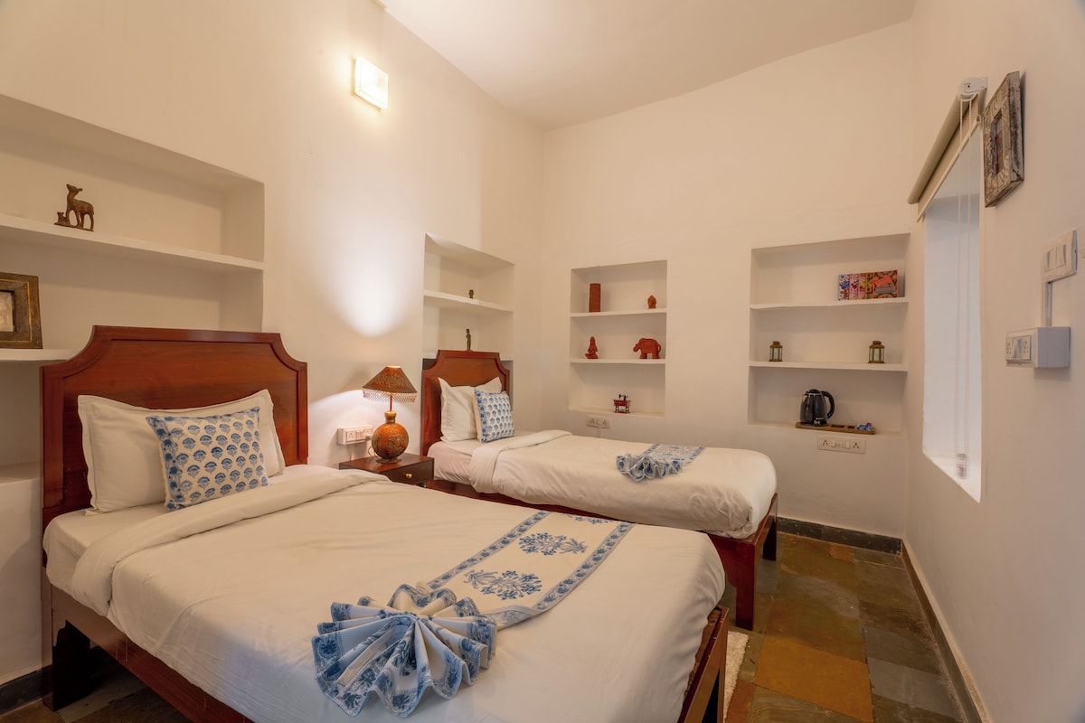 A hotel room with two beds, a nightstand with a lamp, and shelves with decorative items.
