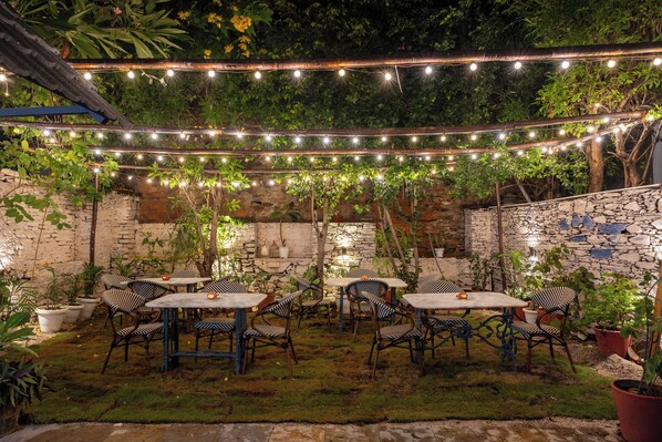 Un espacio al aire libre con mesas, sillas, luces colgantes y plantas en macetas.