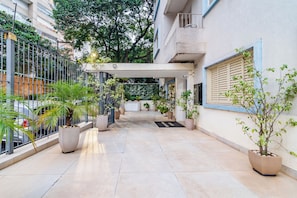Un couloir extérieur abrité, agrémenté de plantes en pot, avec une clôture en métal et l’entrée d’un bâtiment.
