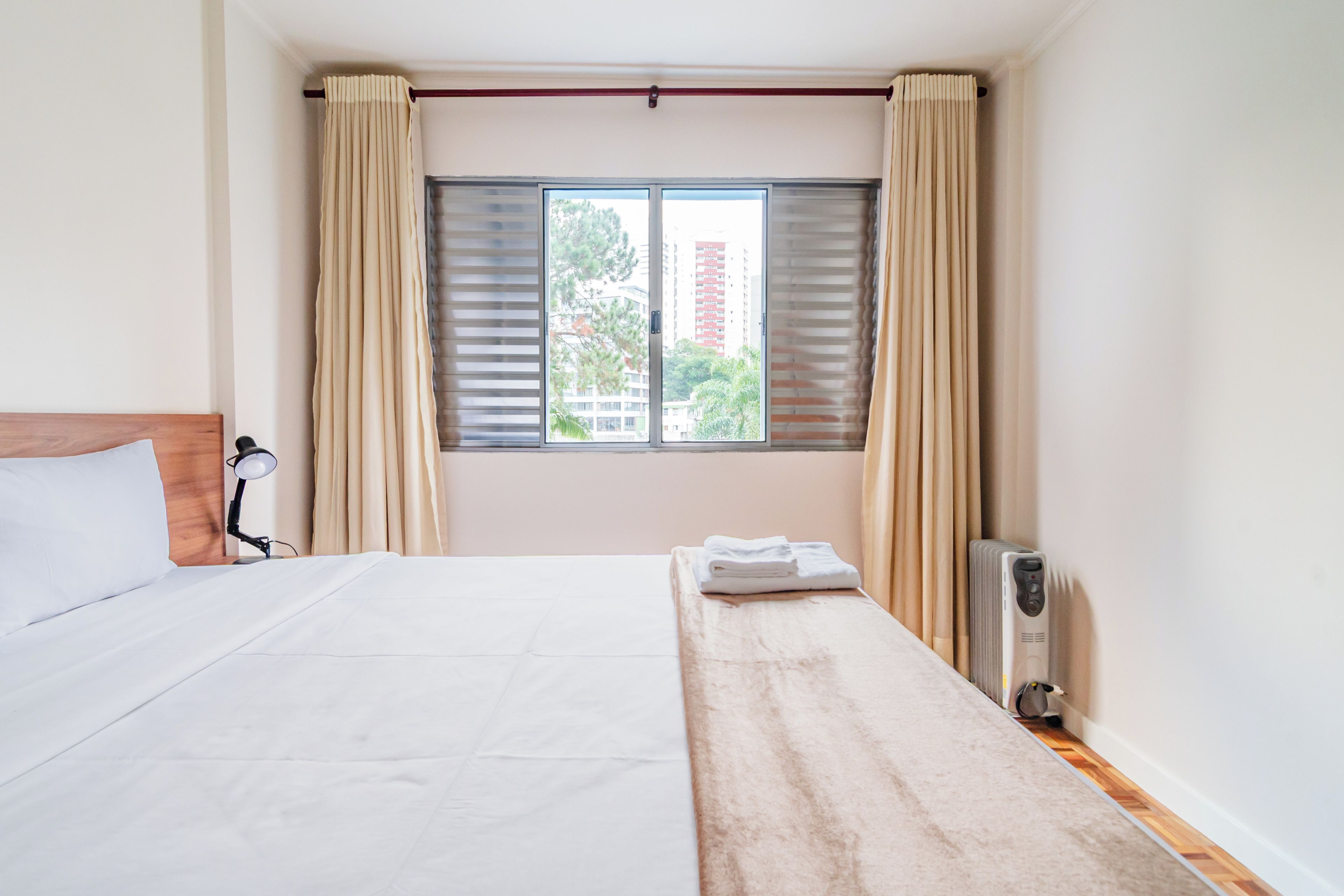 Habitación de hotel con cama, mesita de noche, lámpara, radiador y ventana con cortinas.