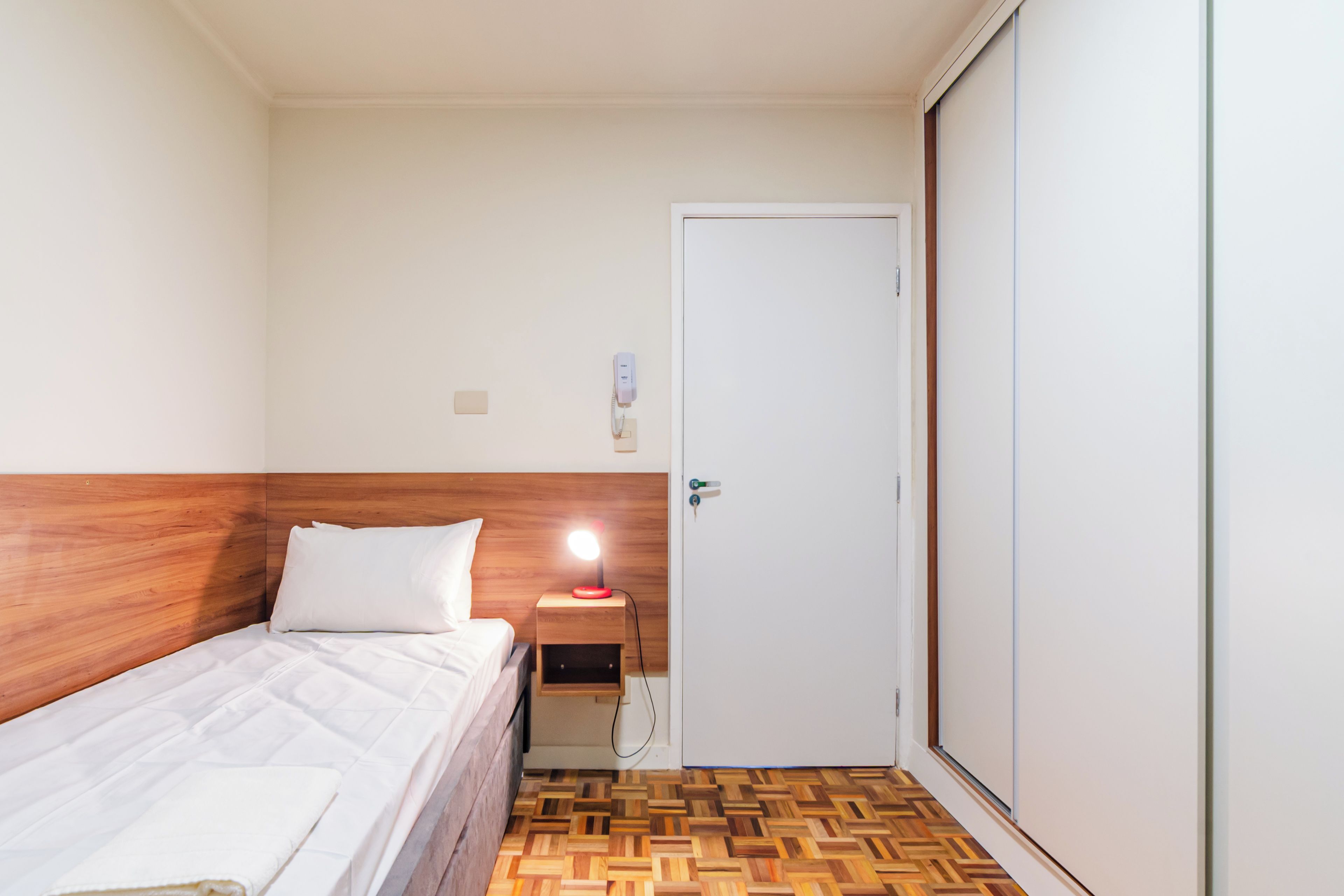 A hotel room with a wooden headboard, a bed with white bedding, a bedside table with a lamp, and a door.