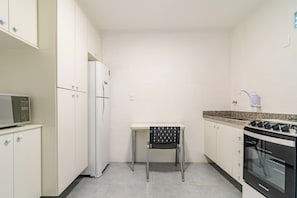 Une cuisine compacte avec des meubles blancs, un four à micro-ondes, un réfrigérateur, une cuisinière à gaz et une petite table à manger avec une chaise.