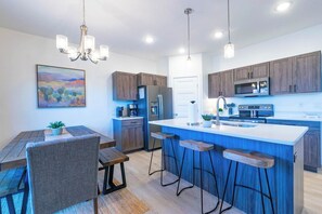 Une cuisine moderne avec des meubles en bois, un îlot central doté de tabourets de bar, une table à manger avec des chaises et des luminaires suspendus.