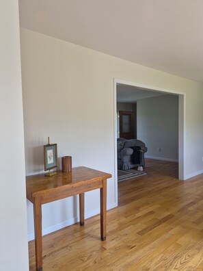 A room with a wooden floor, a small table with a picture frame, and a doorway leading to another room with a patterned sofa. - Peaceful Country 3 Bedroom Home near Martinsburg, Shepherdstown and Hagerstown. (Martinsburg)