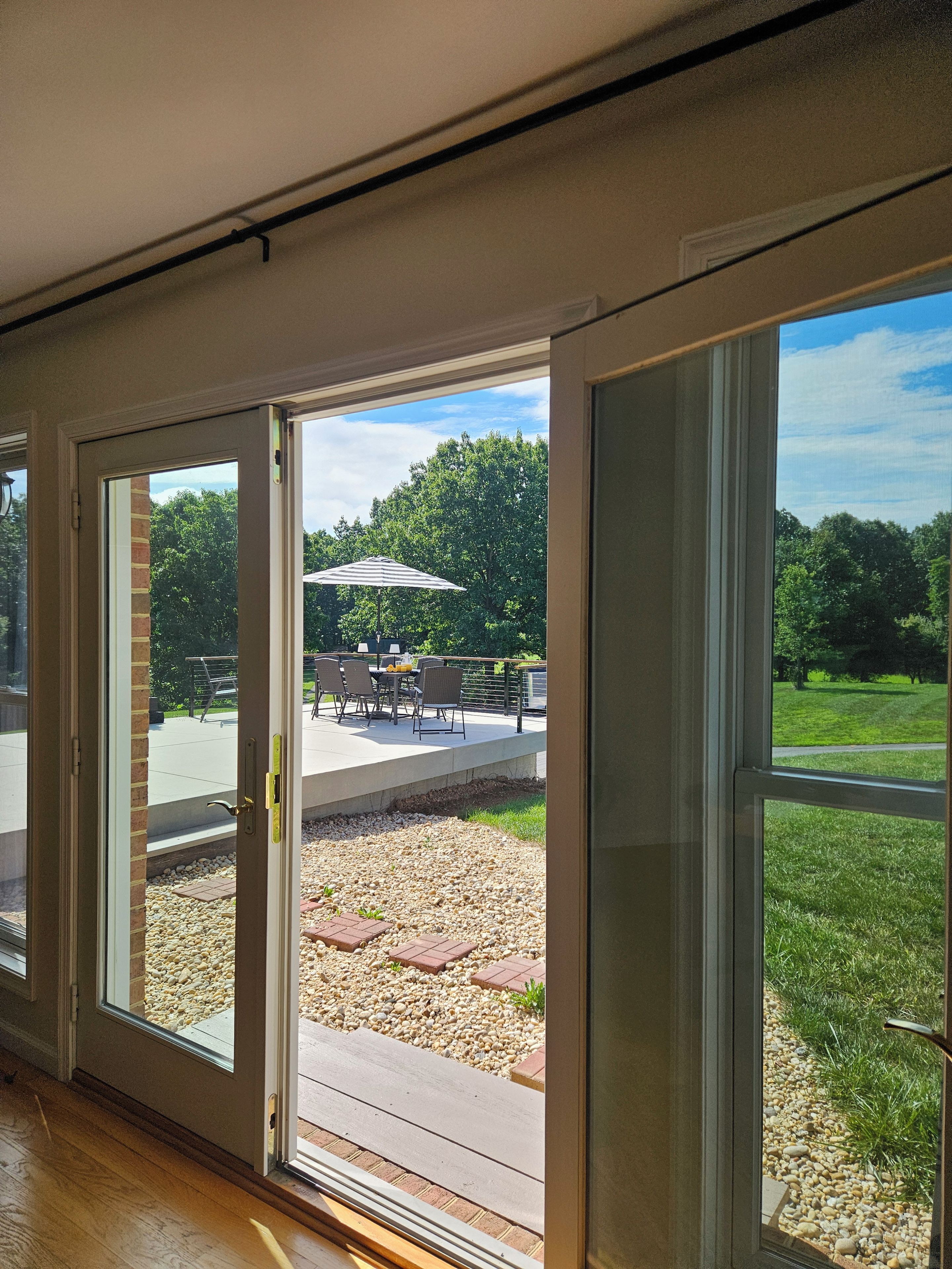 A view through a sliding glass door into an outdoor patio area with a table and chairs, a grassy lawn, and trees in the background.