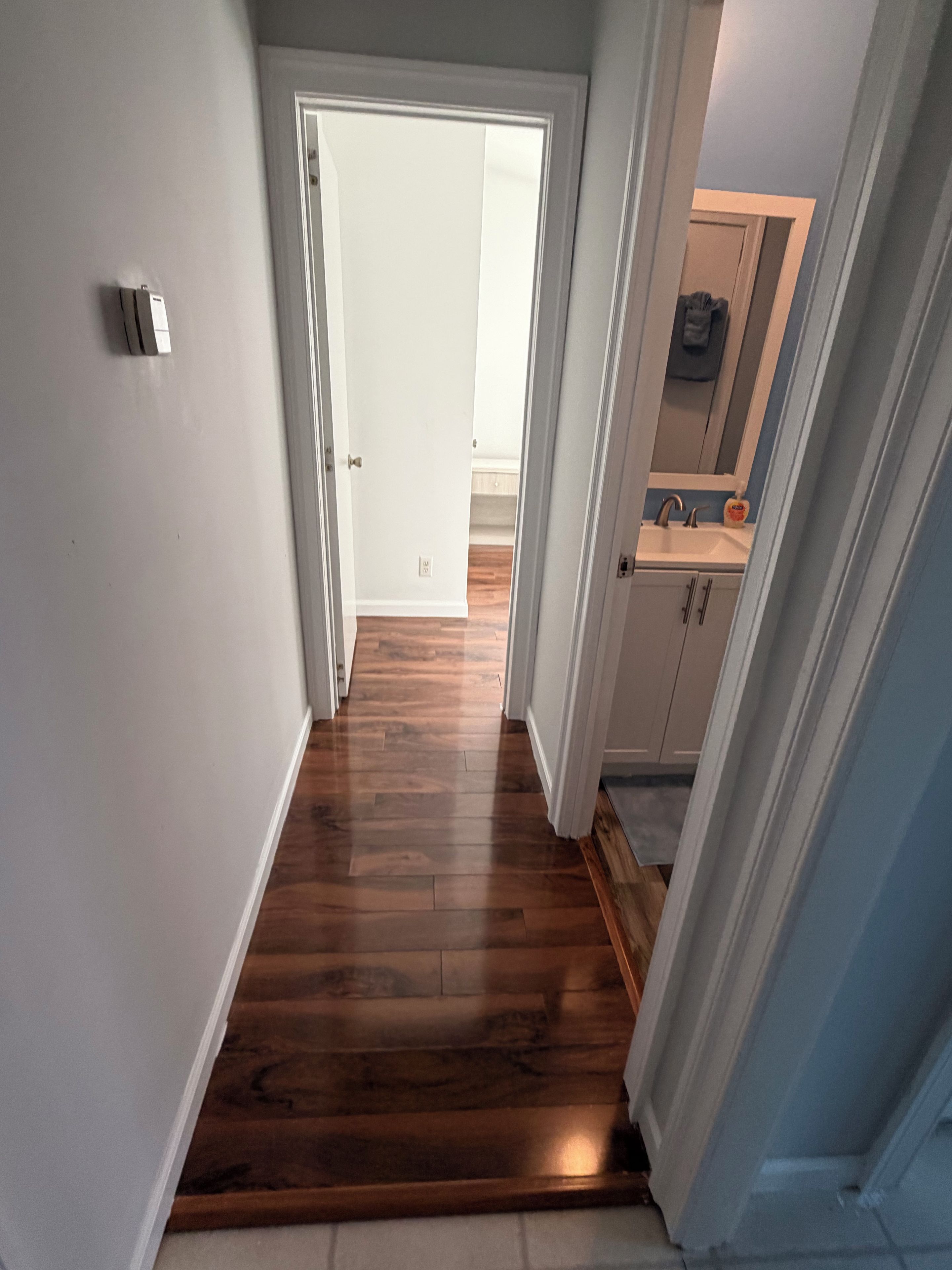 A hallway with wooden flooring leading to a bathroom with a sink and mirror.