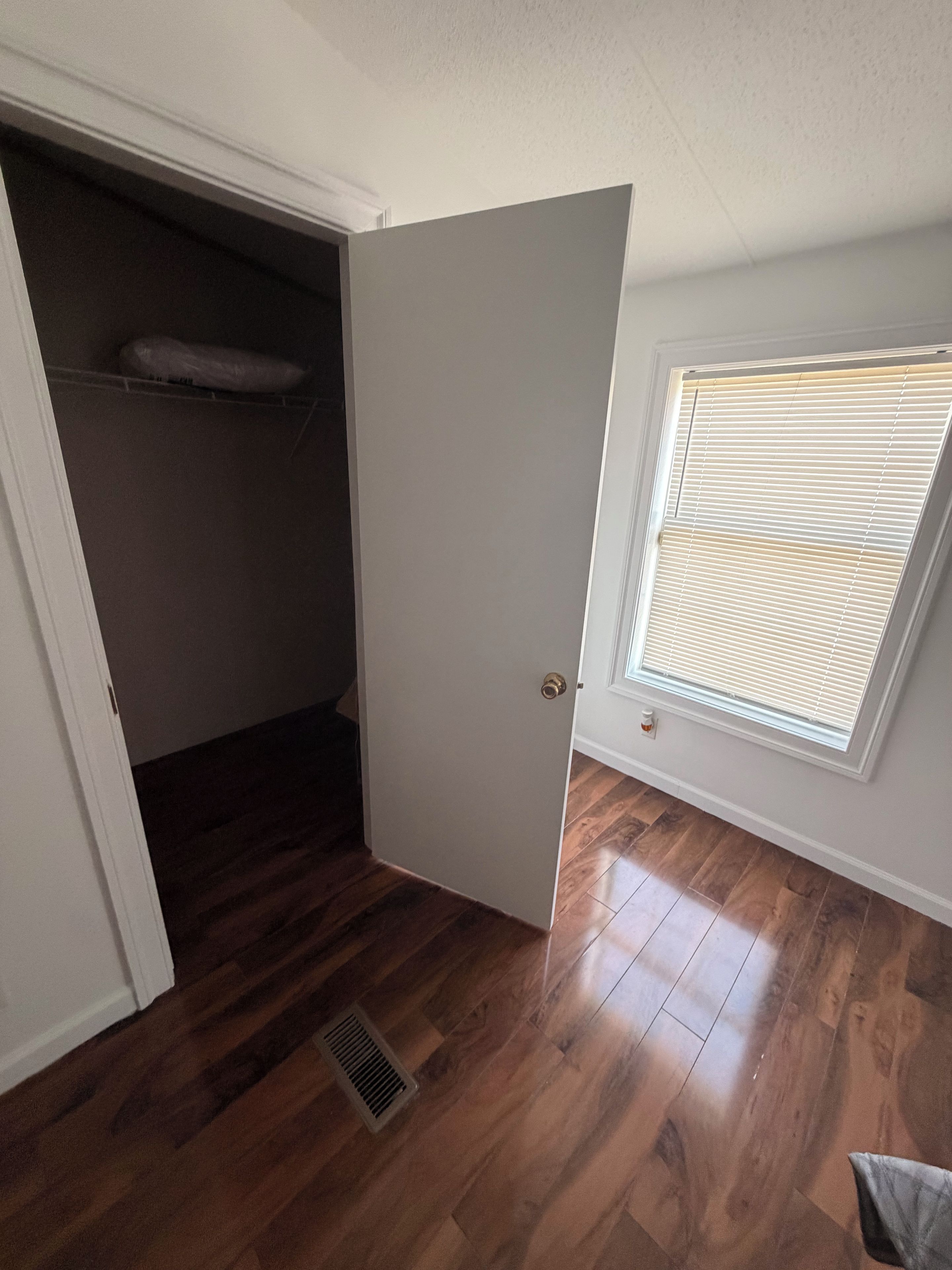 A room with a wooden floor, a white door, a window with blinds, and a vent.