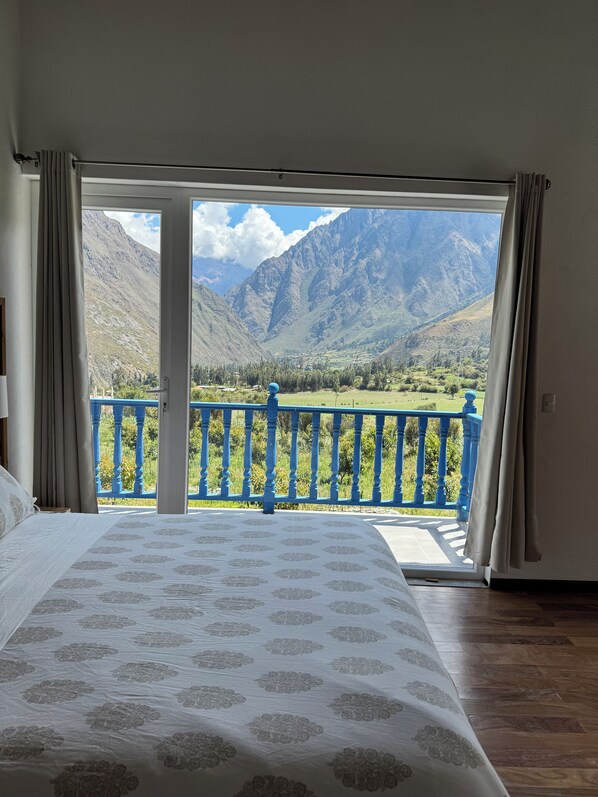 A bedroom with a large bed, a view of mountains through an open door, and a blue balcony railing.