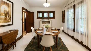 A dining room with a round table set for six, flanked by white chairs, a green patterned rug, and a wooden cabinet.