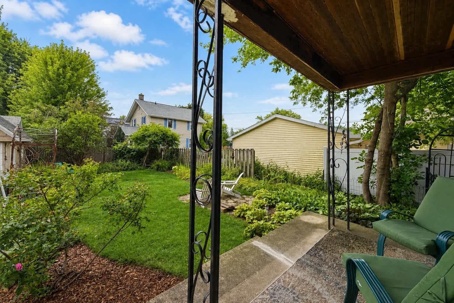 A covered patio area with a green chair, a swing set, and a garden with various plants and a lawn.
