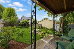 A covered patio area with a green chair, a swing set, and a garden with various plants and a lawn. - Peabody Park Retreat | Sleeps 5 | Fire Pit (Appleton)