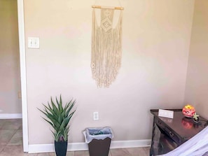 Een kamer met een macramé wanddecoratie, een potplant, een prullenbak en een klein tafeltje met een klok en decoratieve objecten.