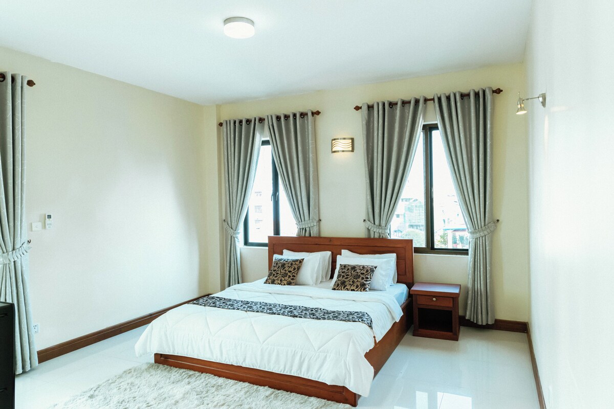 Um quarto com cama de madeira, mesa de cabeceira e uma janela grande com cortinas.