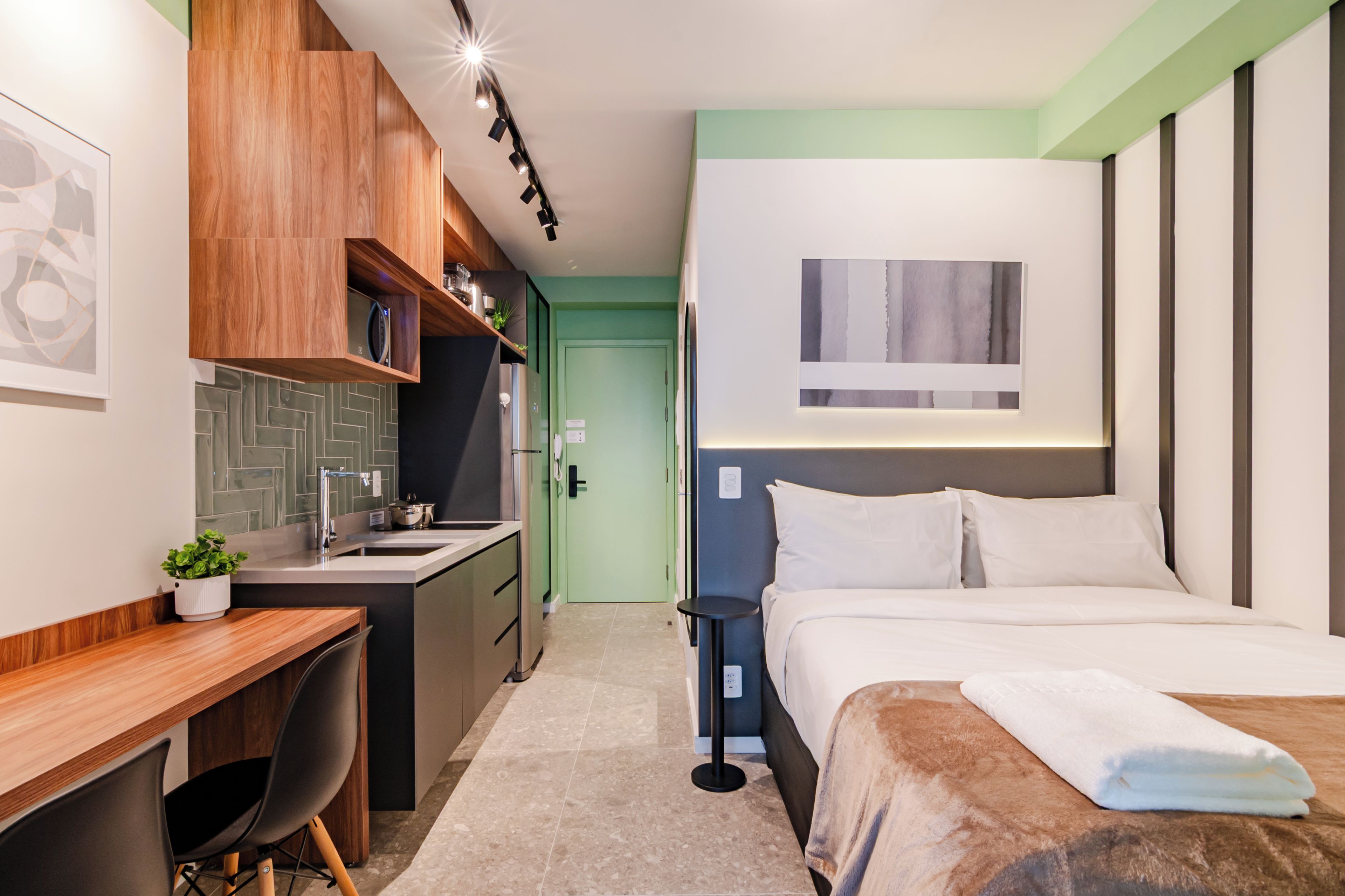 A modern hotel room with a bed, a desk, a kitchenette, and a bathroom.