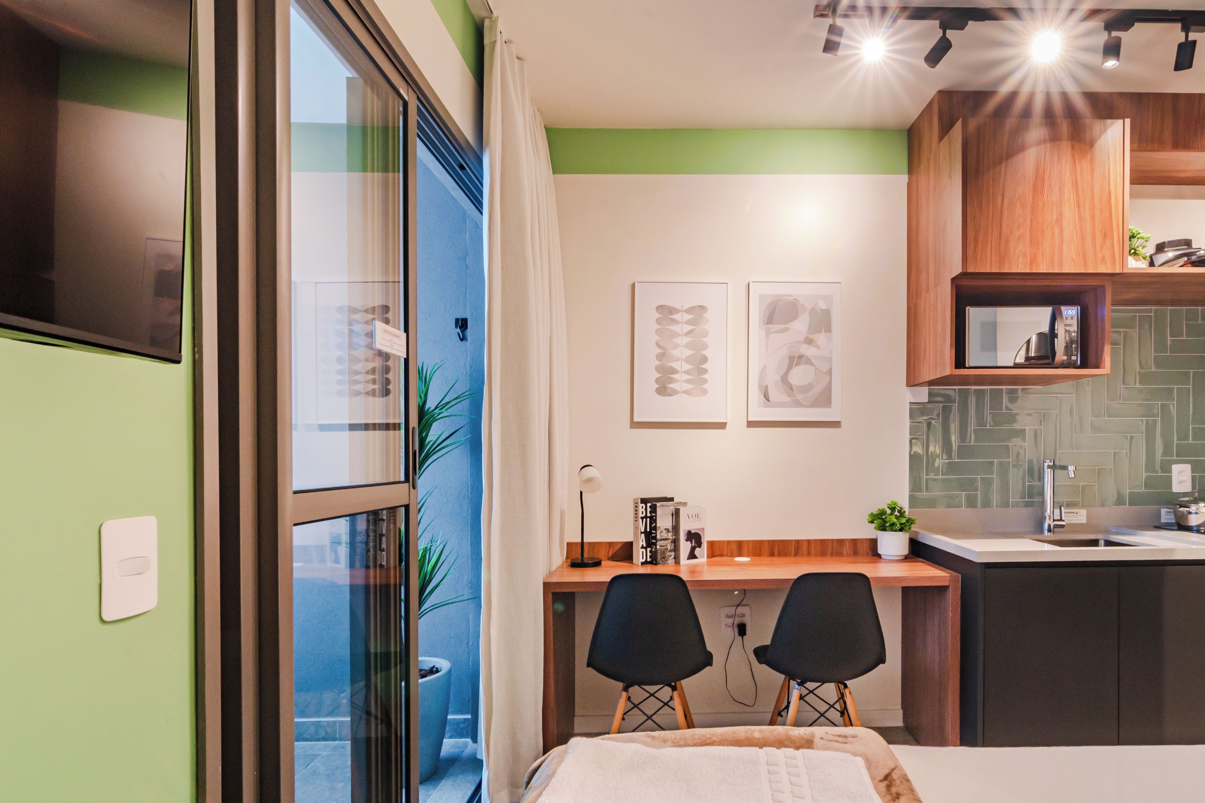 A modern kitchen with a built-in stove, a sink, and a wooden cabinet above the counter. There is a dining area with two chairs and a table, and a bed with a white headboard.