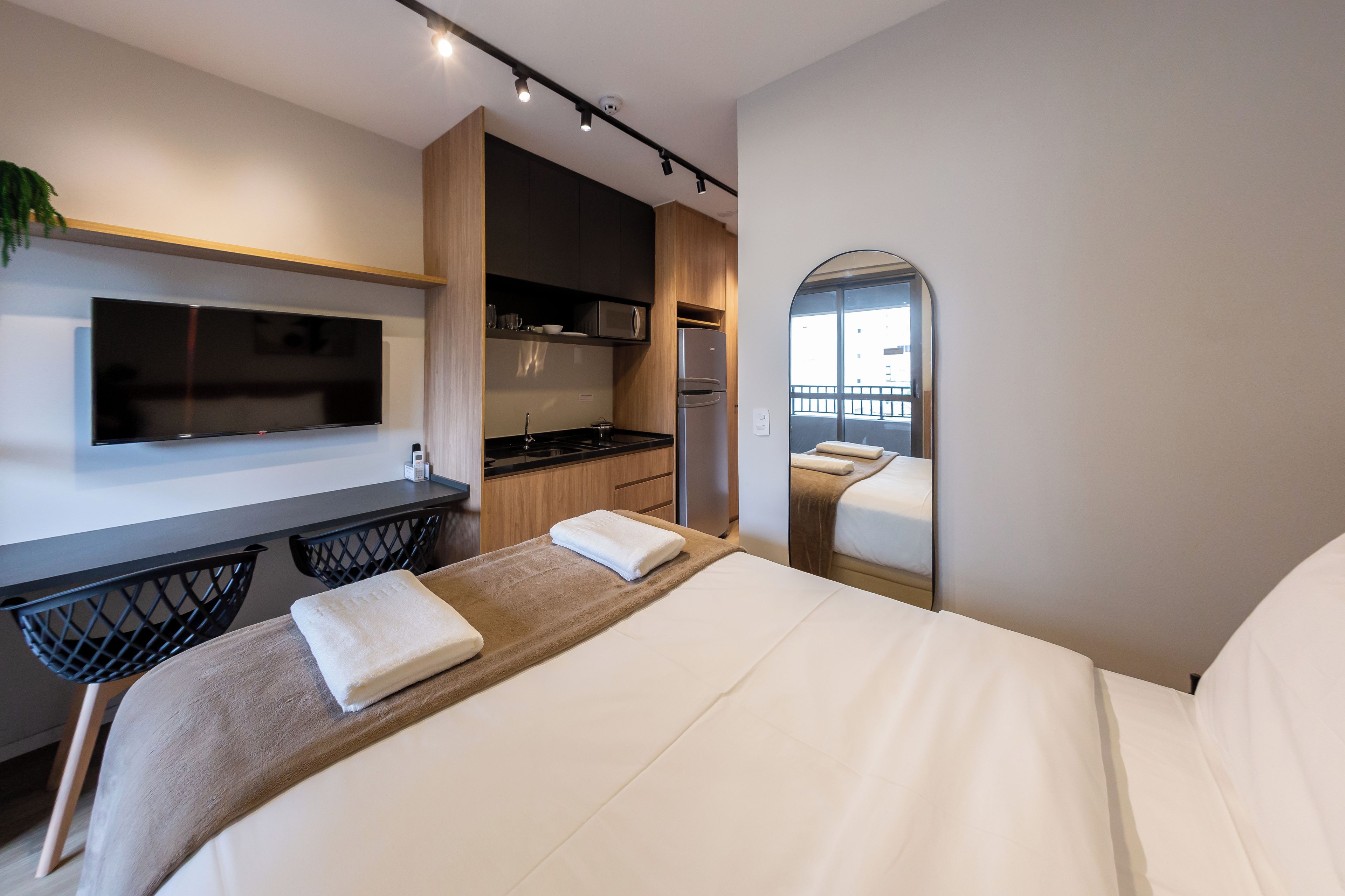 A modern hotel room with a bed, a flat-screen TV, a kitchenette, and a large mirror.
