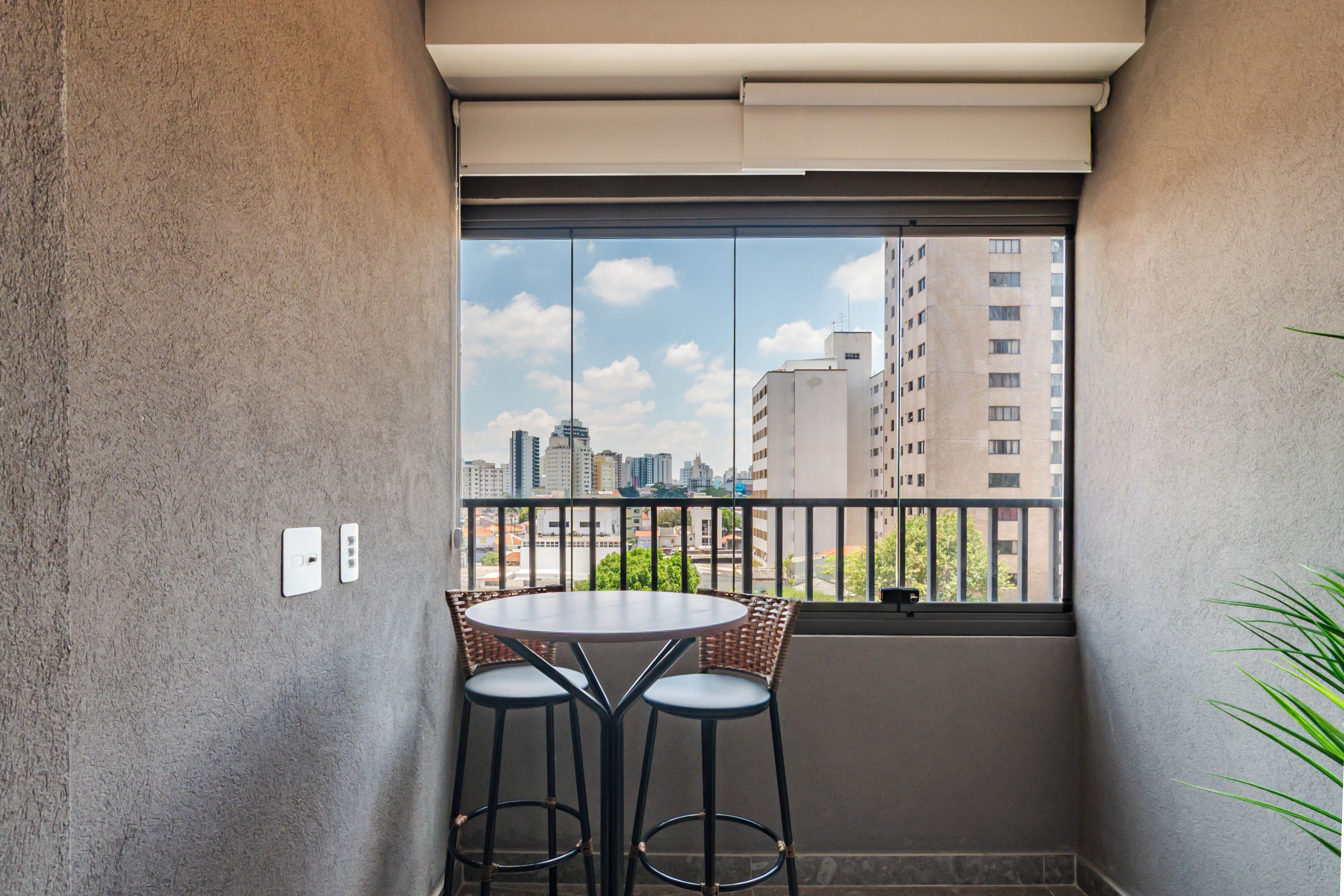 A small balcony with a table and two chairs, a view of a cityscape, and a plant on the right.