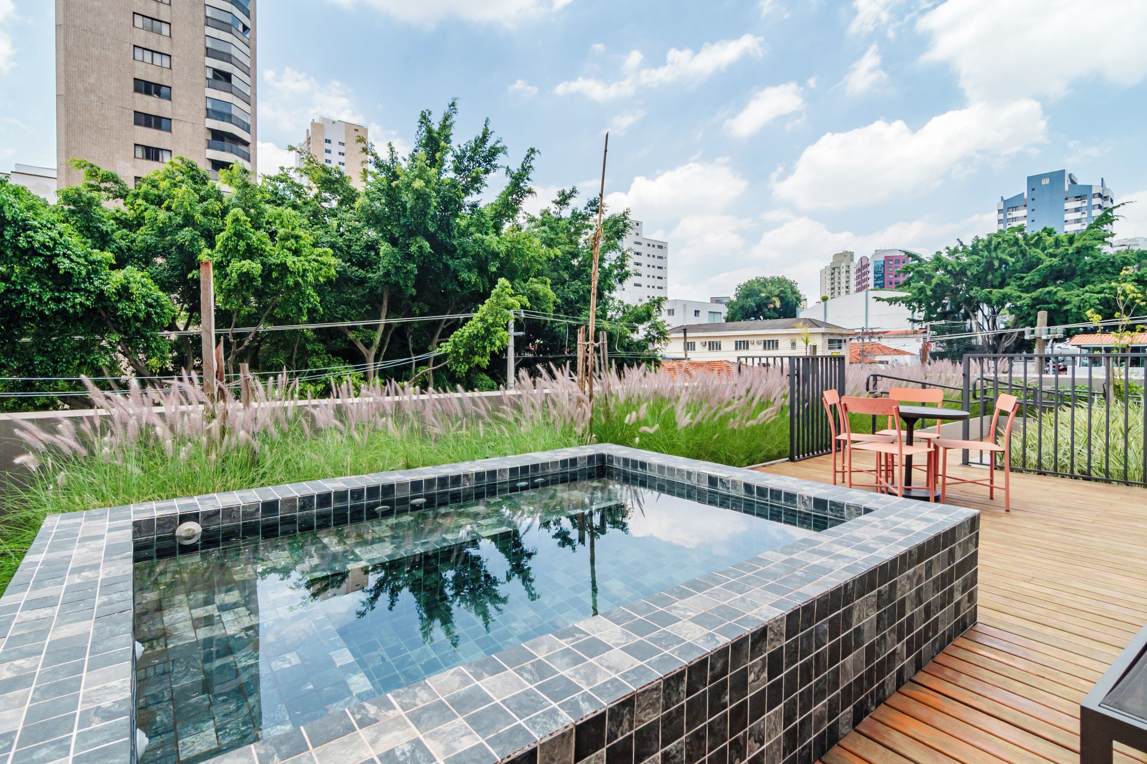 Una piscina en la azotea con cubierta de madera y mobiliario exterior, rodeada de vegetación y edificios de la ciudad.