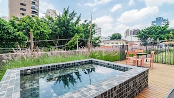 Una piscina en la azotea con cubierta de madera y mobiliario exterior, rodeada de vegetación y edificios de la ciudad.