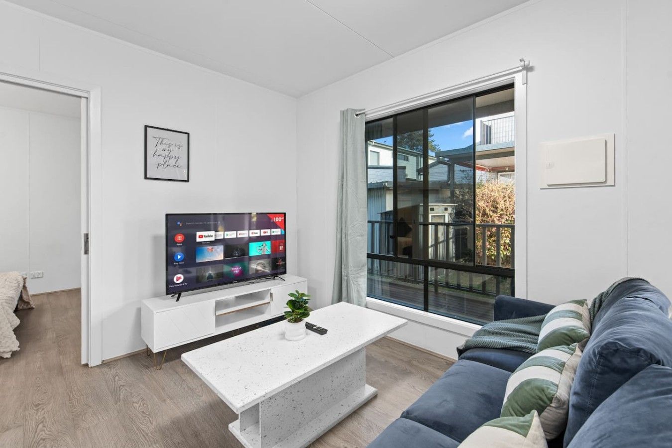 A modern living room with a flat-screen TV, a white coffee table, a sofa, and a large window with a view of a residential area.