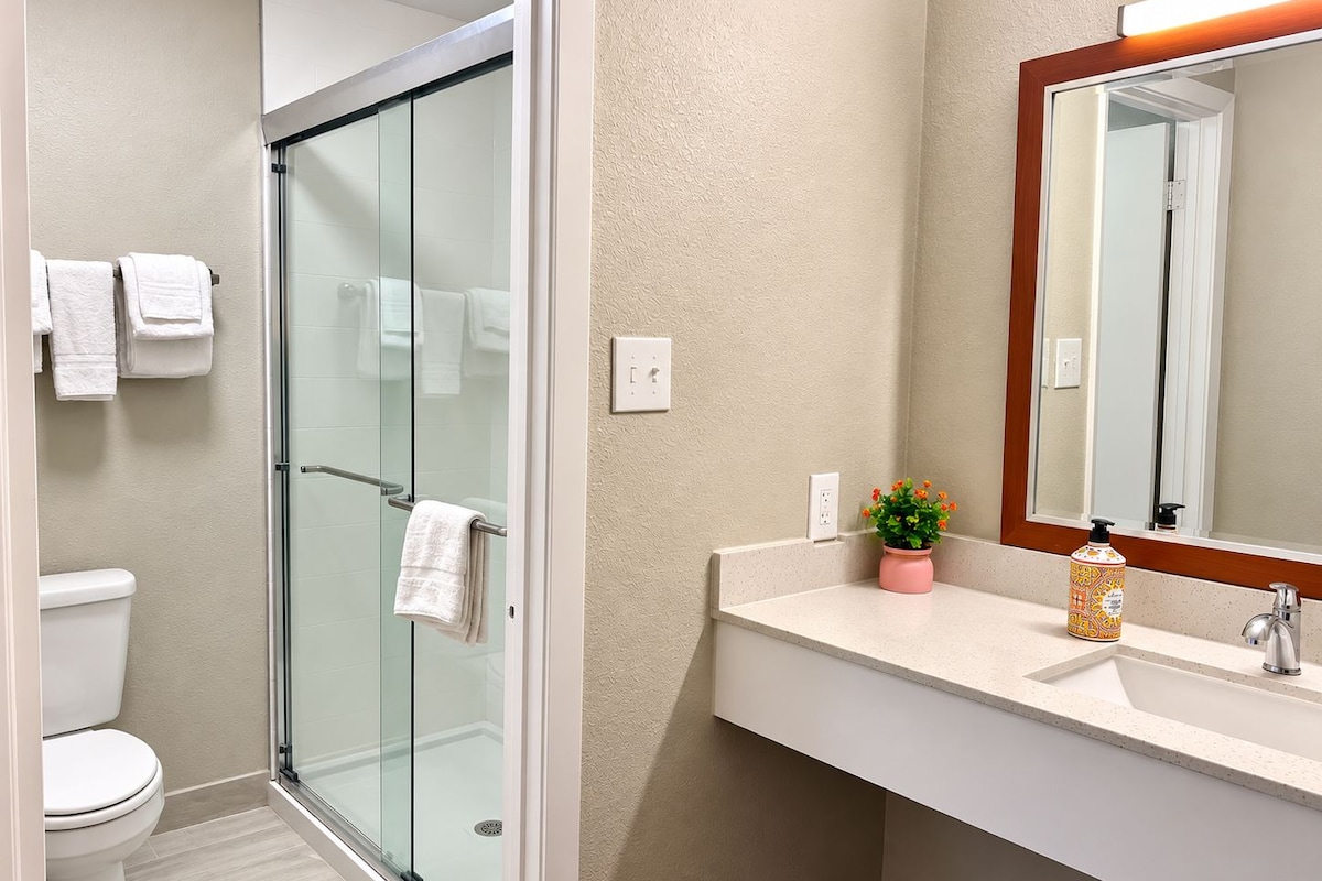 A bathroom with a glass-enclosed shower, a white toilet, a sink with a mirror, and a small potted plant.