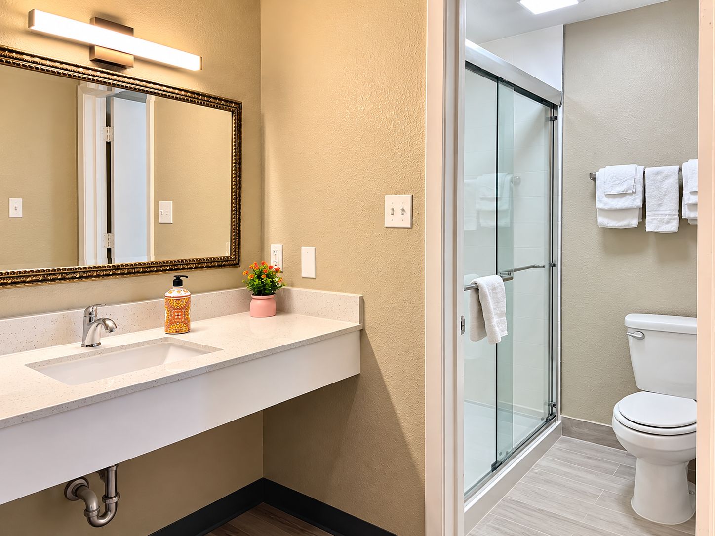 Un baño con cabina de ducha de vidrio, inodoro blanco y lavamanos con espejo y dispensador de jabón.