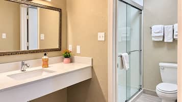 Un baño con cabina de ducha de vidrio, inodoro blanco y lavamanos con espejo y dispensador de jabón.