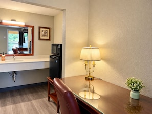A hotel room with a wooden desk, a lamp, a small potted plant, a refrigerator, a microwave, a sink, and a mirror. - Remington Inn and Extended Stay (Little Rock)