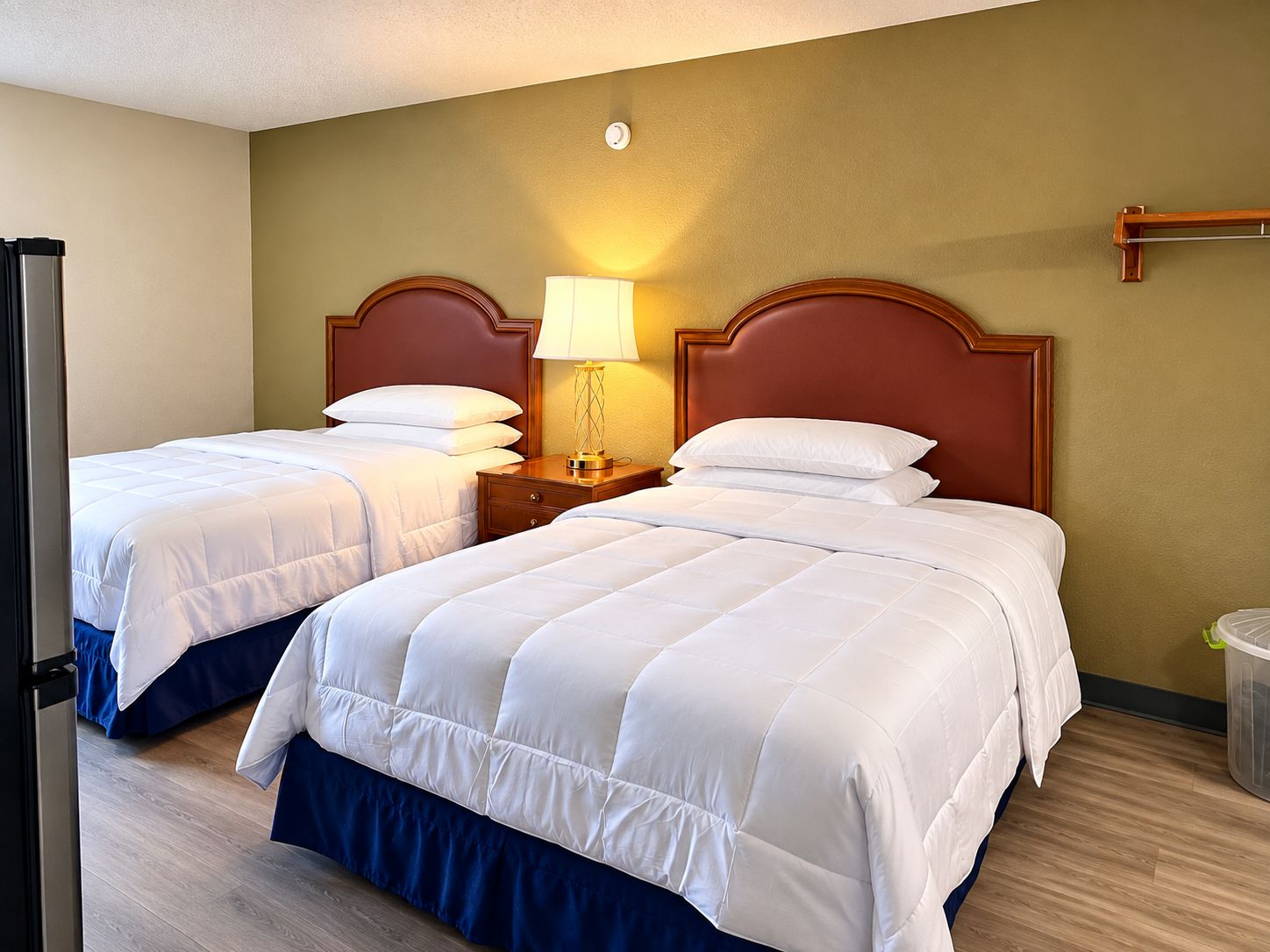 A hotel room with two beds, a nightstand with a lamp, and a trash can.
