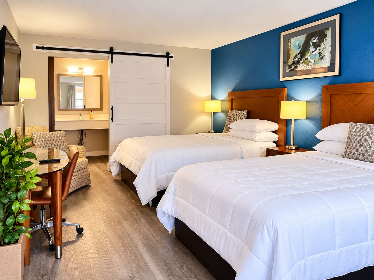 Habitación de hotel con dos camas, un escritorio con silla, un espejo y un cuadro en la pared.