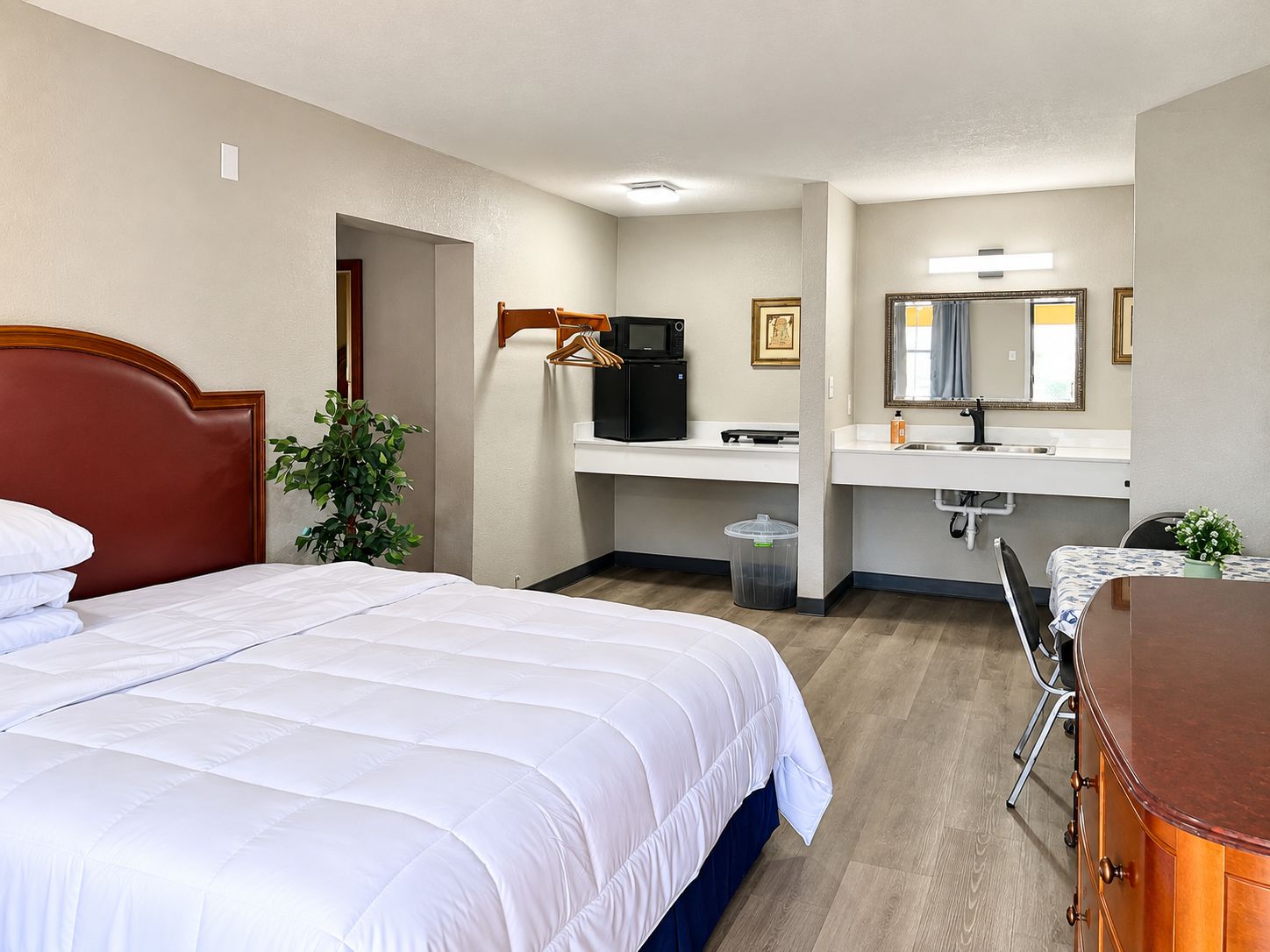 A hotel room with a bed, a desk, a chair, a mirror, a sink, and a trash bin.