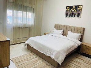 A bedroom with a bed, a nightstand, a wooden dresser, a window with sheer curtains, and a wall-mounted artwork. - 3-bedroom apartment in soothing Sousse with AC for relaxing stays (Susah)