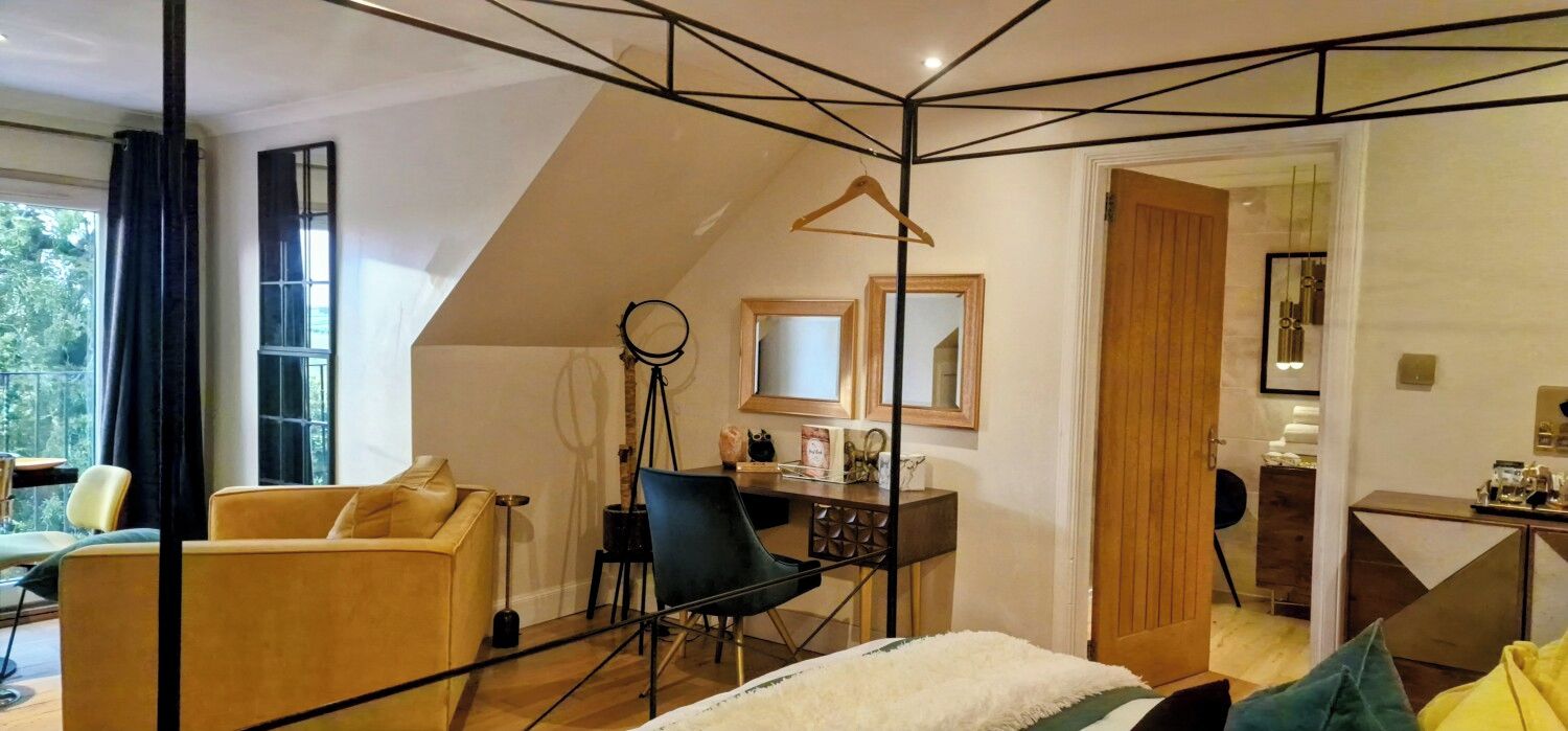 Ett modernt rum med en säng på ett loft, ett skrivbord och en stol. Det finns ett stort fönster som släpper in naturligt ljus och ger utsikt över grönområden utanför.