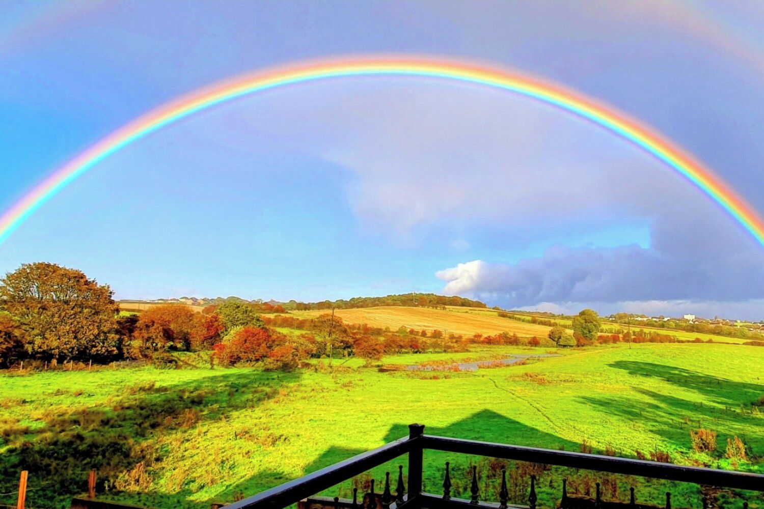Un paysage avec un arc-en-ciel s’étendant au-dessus d’un champ verdoyant et une clôture en bois au premier plan.
