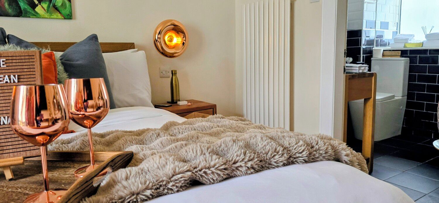 A hotel room with a bed, a nightstand, a wall-mounted light, a fluffy rug, and a wooden side table with a wine glass and a bottle.