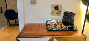 A room with a wooden table, a chair, a coffee machine, and a wall-mounted control panel with various icons.