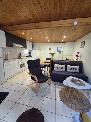 A modern kitchen with white cabinets, a black chair, a dining table, and a sofa with floral cushions. - A cozy little nest in Mont-Dore (Mont-Dore)