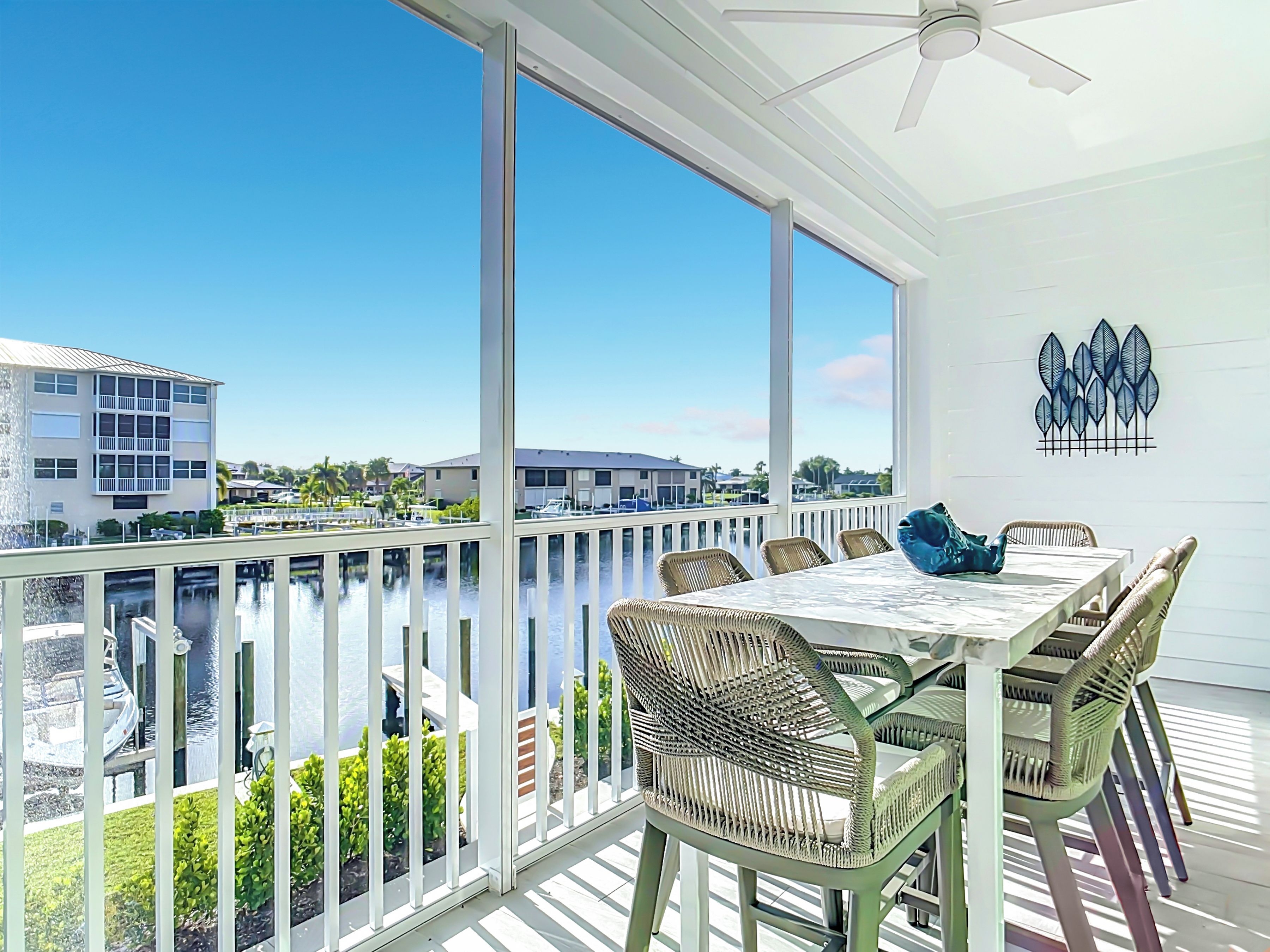 A balcony with a view of a marina and buildings, a table with chairs, and a ceiling fan.