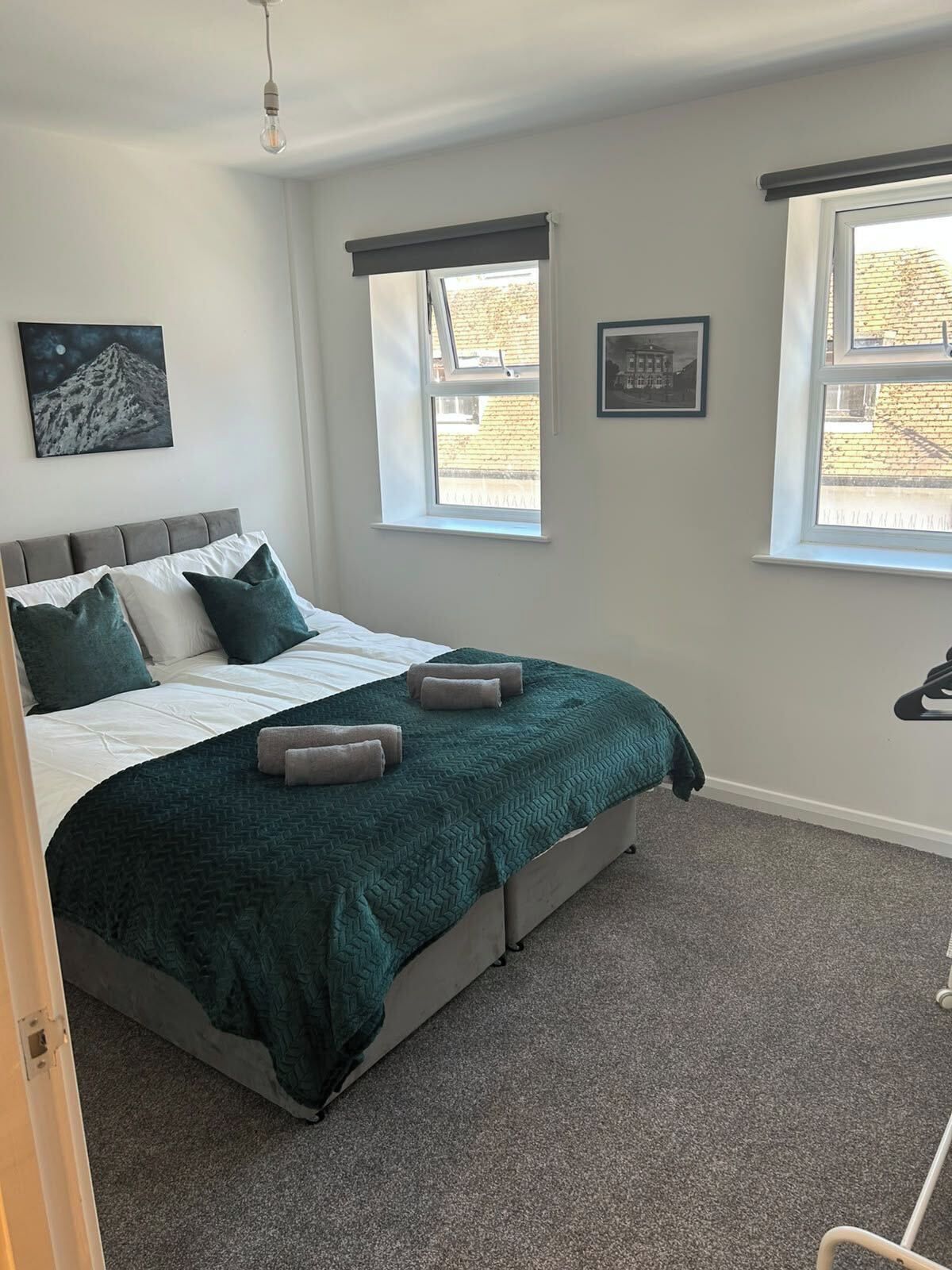 A bedroom with a bed, two windows, a picture on the wall, and a light fixture.