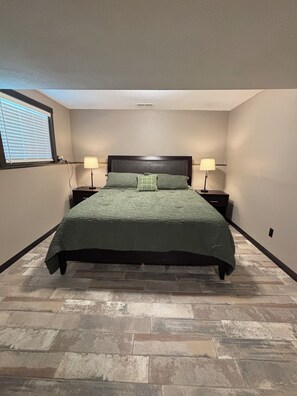 Un dormitorio con una cama grande, dos mesitas de noche con lámparas, una ventana con persianas y un suelo con diseño de piedra.