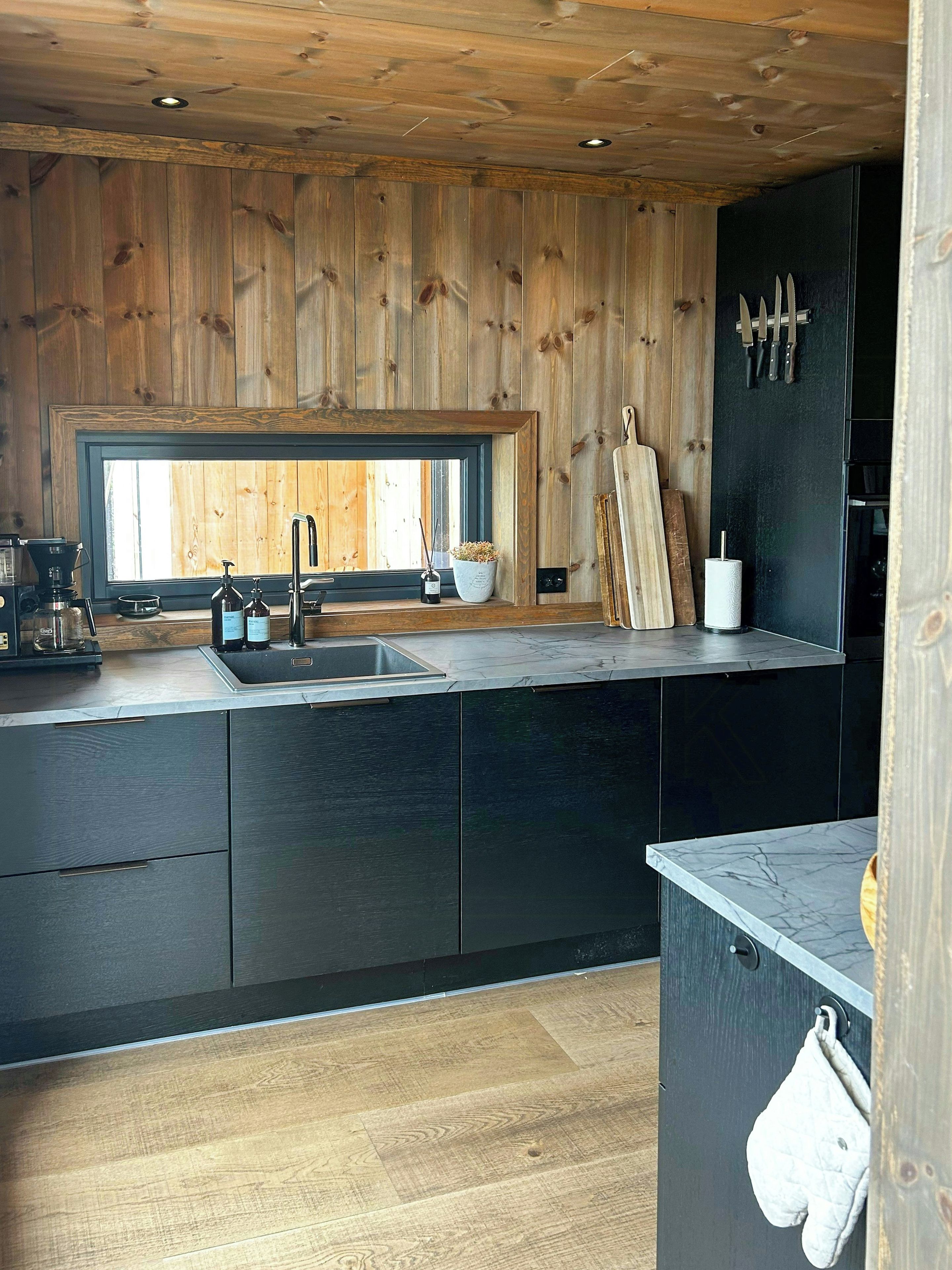 Eine moderne Küche mit Holzvertäfelung, einem großen Fenster und einer schwarzen Arbeitsplatte.