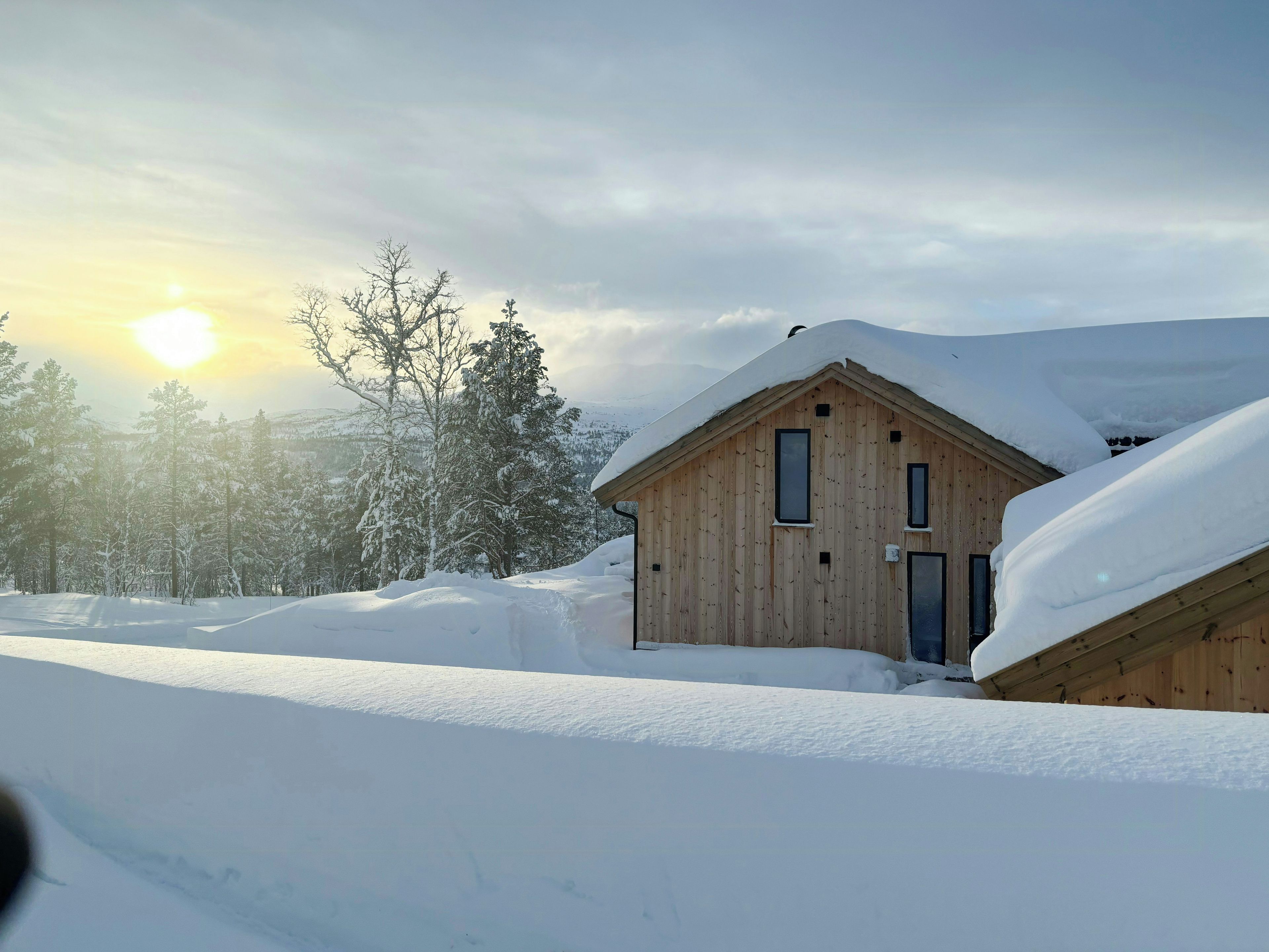 A wooden cabin with a sloped roof covered in snow, surrounded by a snowy landscape and trees.
