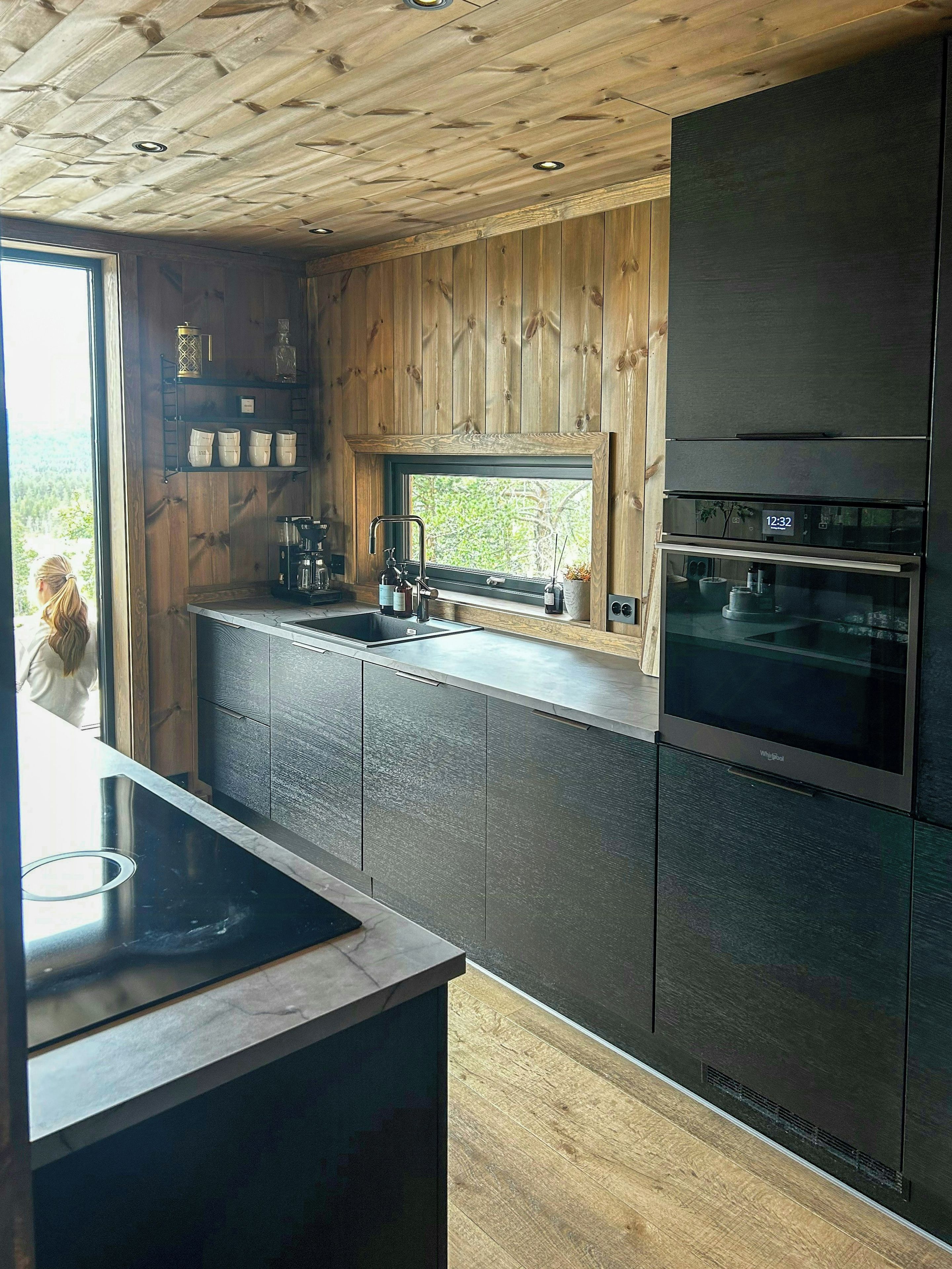 A modern kitchen with a wooden ceiling, built-in appliances, and a window overlooking greenery.